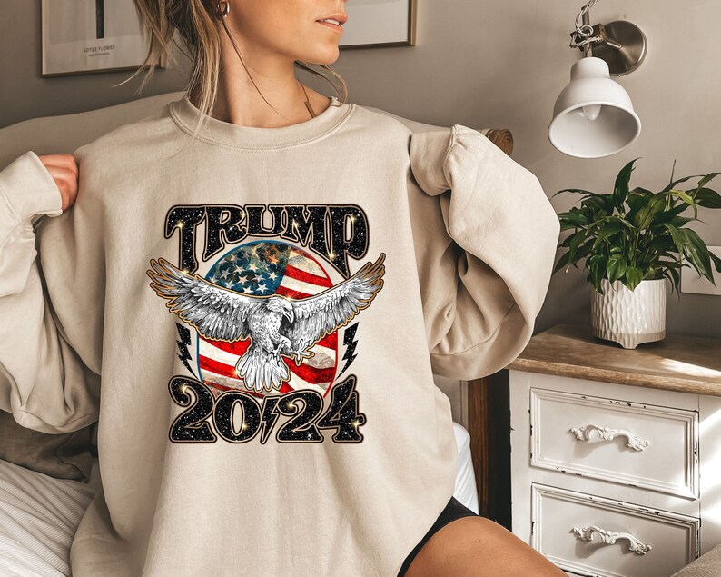 May include: A beige sweatshirt with a graphic print of an eagle flying over an American flag with the text "TRUMP 2024" in black.