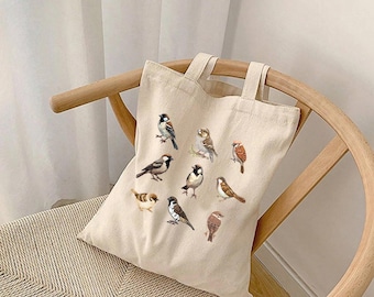 May include: A beige canvas tote bag featuring a collection of illustrated birds in various poses. The birds are depicted in shades of brown, gray, and white, with detailed feather patterns. The bag has two handles and is resting on a wooden chair.