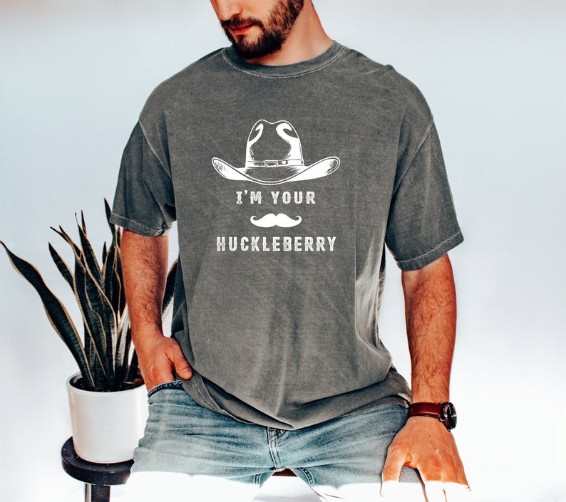 Puede incluir: Camiseta gris con el texto blanco "I'M YOUR HUCKLEBERRY", un sombrero de vaquero y un bigote. La camiseta tiene un corte holgado y mangas cortas.