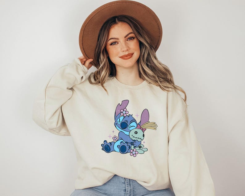 K&ouml;nnte beinhalten: Cremefarbenes Sweatshirt mit einer Cartoonfigur in Blau-, Lila- und Rosat&ouml;nen. Die Grafik zeigt die Figur, die eine Puppe umarmt, umgeben von Blumen. Das Sweatshirt hat lange &Auml;rmel und einen Rundhalsausschnitt.