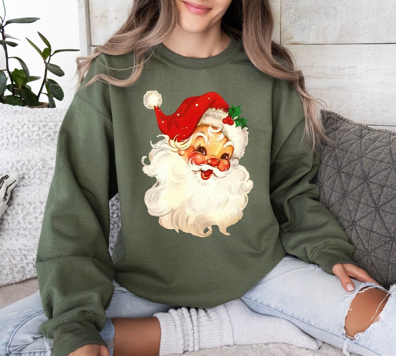 May include: A green sweatshirt with a vintage illustration of Santa Claus's face. The illustration features a red hat with white trim and a white beard.