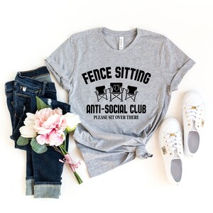 May include: A grey t-shirt with the text "Fence Sitting Anti-Social Club Please Sit Over There" and a graphic of three chairs. The shirt is laid out on a white background with a pair of white trainers, a bouquet of pink flowers, and a pair of blue jeans.