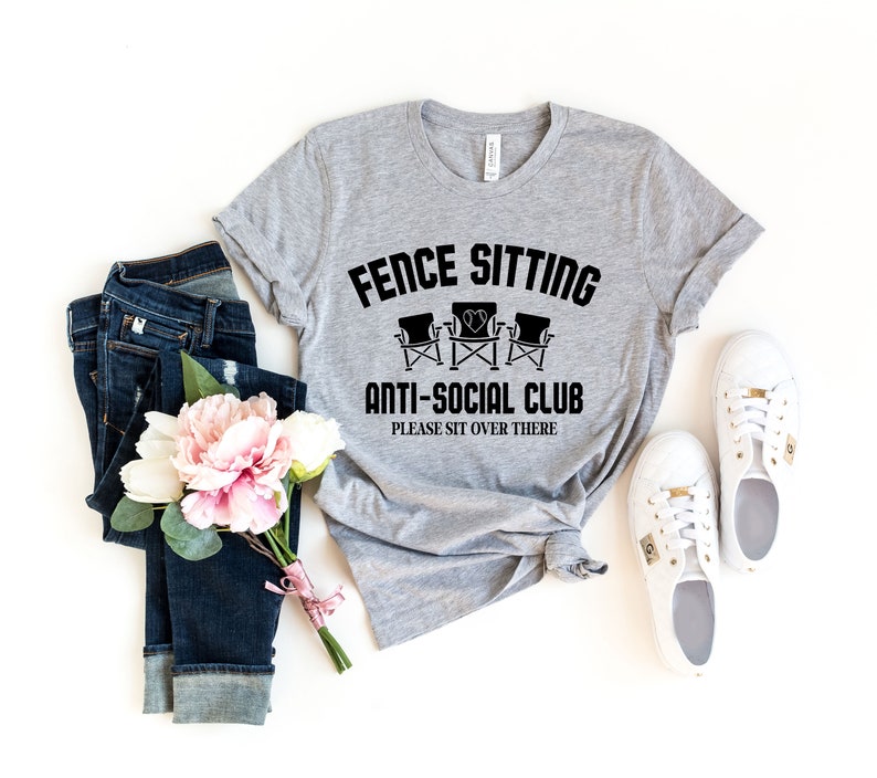 May include: A grey t-shirt with the text "Fence Sitting Anti-Social Club Please Sit Over There" and a graphic of three chairs. The shirt is laid out on a white background with a pair of white trainers, a bouquet of pink flowers, and a pair of blue jeans.