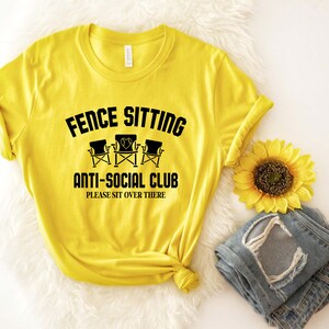 May include: Yellow t-shirt with black text that reads "Fence Sitting Anti-Social Club Please Sit Over There". The text is surrounded by a black circle. There are three black folding chairs with hearts on the back of the chairs.