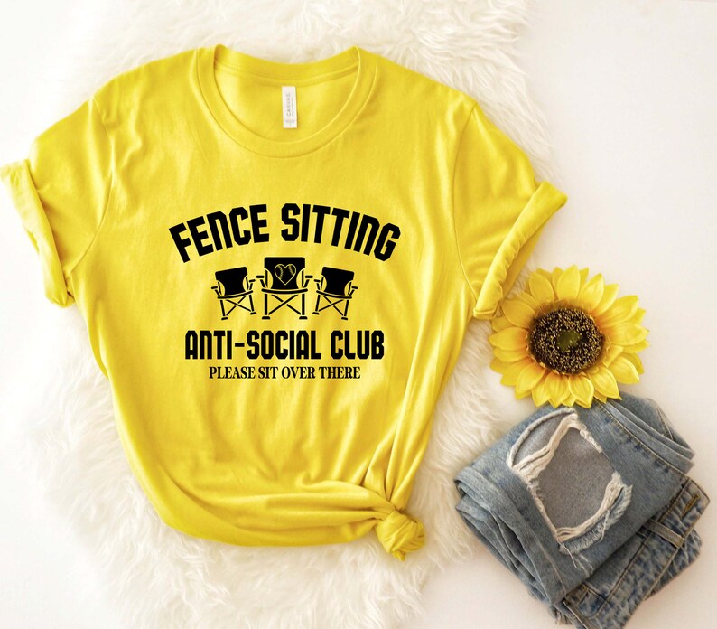 May include: Yellow t-shirt with black text that reads "Fence Sitting Anti-Social Club Please Sit Over There". The text is surrounded by a black circle. There are three black folding chairs with hearts on the back of the chairs.