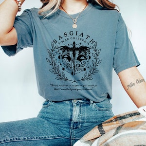 Peut inclure: Un t-shirt bleu avec un motif graphique noir repr&eacute;sentant un dragon, une couronne de laurier et le texte "BASGIATH WAR COLLEGE". Le texte "There's nowhere in existence you could go that I wouldn't find you, Violence." est &eacute;galement imprim&eacute; sur le t-shirt.