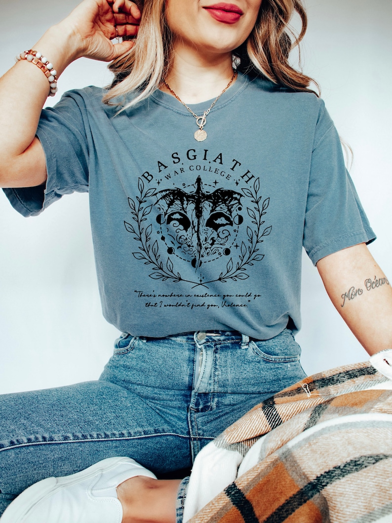 Peut inclure: Un t-shirt bleu avec un motif graphique noir repr&eacute;sentant un dragon, une couronne de laurier et le texte "BASGIATH WAR COLLEGE". Le texte "There's nowhere in existence you could go that I wouldn't find you, Violence." est &eacute;galement imprim&eacute; sur le t-shirt.