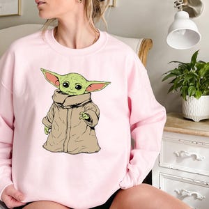 May include: A pink sweatshirt with a graphic of a green alien character wearing a brown robe. The character has large ears and big eyes.