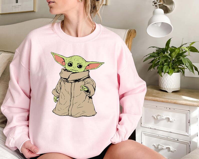 May include: A pink sweatshirt with a graphic of a green alien character wearing a brown robe. The character has large ears and big eyes.