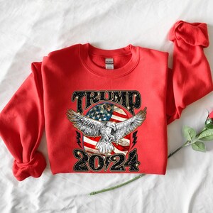 May include: Red sweatshirt with a graphic of an eagle flying over an American flag with the text "TRUMP 2024" in black glitter.