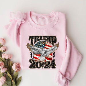 May include: Pink sweatshirt with a graphic design of an eagle flying over an American flag with the text "TRUMP 2024" in black.