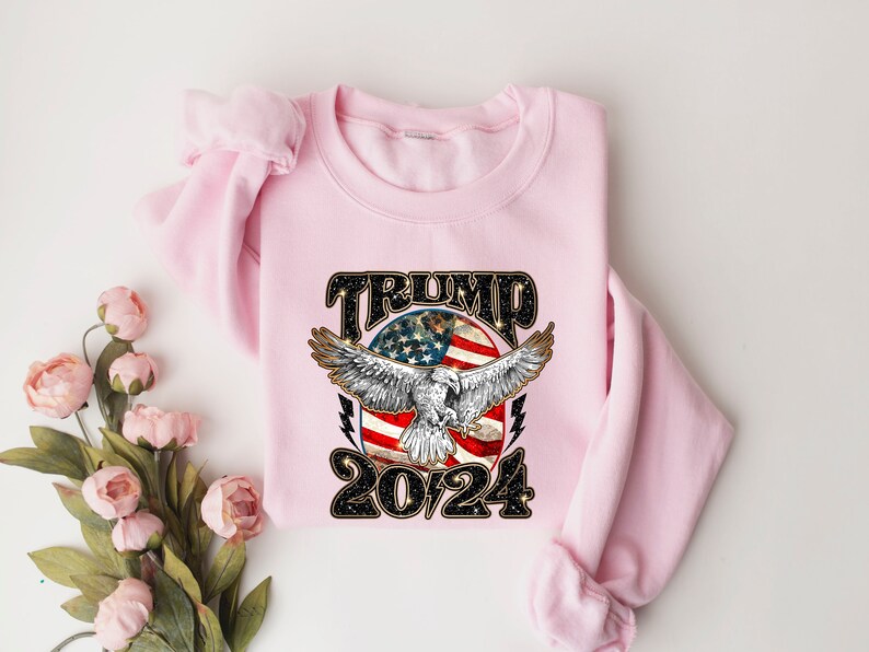 May include: Pink sweatshirt with a graphic design of an eagle flying over an American flag with the text "TRUMP 2024" in black.