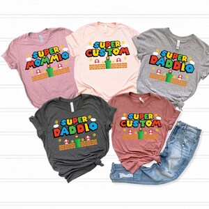 May include: Five t-shirts in various colors, each featuring a Super Mario-themed design. The shirts display the words "Super Mommio", "Super Custom", and "Super Daddio" in a colorful, playful font, with accompanying graphics.