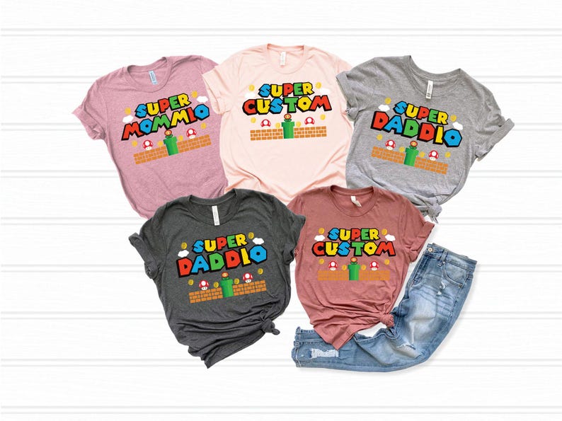 May include: Five t-shirts in various colors, each featuring a Super Mario-themed design. The shirts display the words "Super Mommio", "Super Custom", and "Super Daddio" in a colorful, playful font, with accompanying graphics.