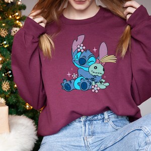 K&ouml;nnte beinhalten: Burgunderfarbenes Sweatshirt mit einer blauen Cartoonfigur, die eine gr&uuml;ne Puppe umarmt. Die Figur hat gro&szlig;e Ohren und ist mit Blumen verziert. Das Sweatshirt wird mit einer blauen Jeans getragen. Im Hintergrund ist ein Weihnachtsbaum zu sehen.