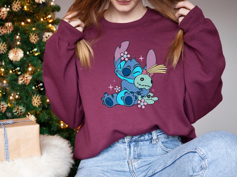 K&ouml;nnte beinhalten: Burgunderfarbenes Sweatshirt mit einer blauen Cartoonfigur, die eine gr&uuml;ne Puppe umarmt. Die Figur hat gro&szlig;e Ohren und ist mit Blumen verziert. Das Sweatshirt wird mit einer blauen Jeans getragen. Im Hintergrund ist ein Weihnachtsbaum zu sehen.