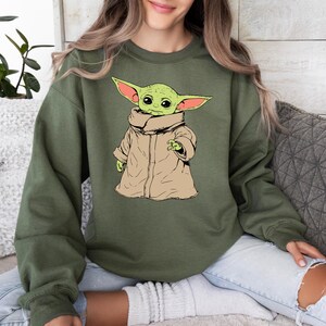 May include: A green sweatshirt with a cartoon image of a green alien wearing a brown coat. The alien has large ears and big eyes.