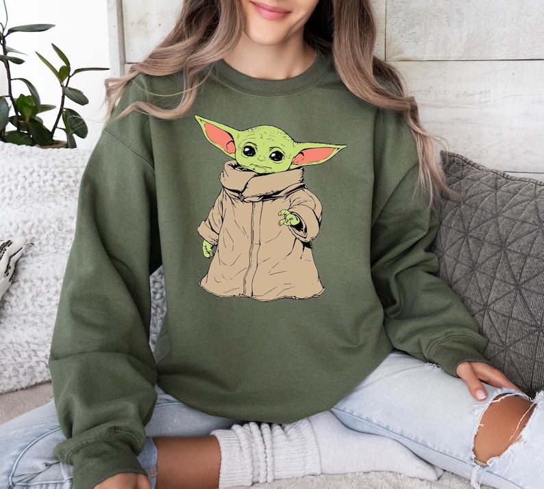 May include: A green sweatshirt with a cartoon image of a green alien wearing a brown coat. The alien has large ears and big eyes.