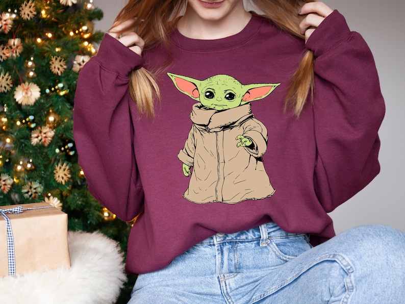 May include: A maroon sweatshirt with a green and brown cartoon character wearing a brown coat. The character has large ears and is looking to the right.