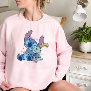 K&ouml;nnte beinhalten: Ein hellrosa Sweatshirt mit einer Cartoonfigur in Blau und Lila, die eine Puppe h&auml;lt. Die Figur ist von kleinen rosa Blumen und Glitzer umgeben. Das Sweatshirt hat eine einfarbige, hellrosa Farbe.