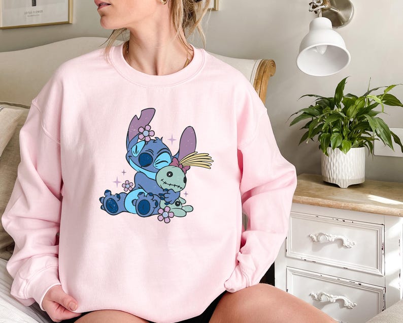 K&ouml;nnte beinhalten: Ein hellrosa Sweatshirt mit einer Cartoonfigur in Blau und Lila, die eine Puppe h&auml;lt. Die Figur ist von kleinen rosa Blumen und Glitzer umgeben. Das Sweatshirt hat eine einfarbige, hellrosa Farbe.
