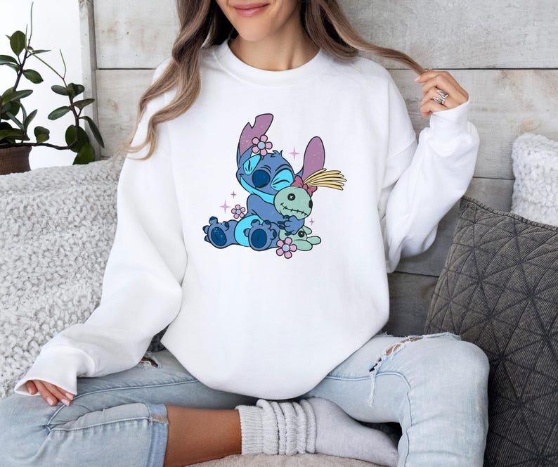 K&ouml;nnte beinhalten: Wei&szlig;es Sweatshirt mit einem Cartoon-Bild von Stitch aus Lilo & Stitch. Die blaue Figur umarmt eine Puppe, umgeben von rosa Blumen und Glitzer. Das Sweatshirt hat lange &Auml;rmel und einen Rundhalsausschnitt.