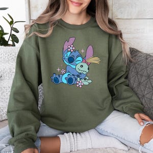 K&ouml;nnte beinhalten: Olivgr&uuml;nes Sweatshirt mit einer Cartoon-Illustration von Stitch, der eine Puppe umarmt. Die Grafik ist in Blaut&ouml;nen gehalten, mit rosa und gelben Akzenten. Das Sweatshirt hat lange &Auml;rmel und einen Rundhalsausschnitt.
