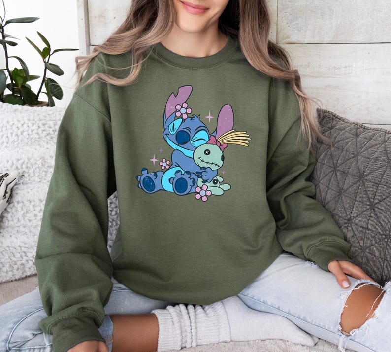 K&ouml;nnte beinhalten: Olivgr&uuml;nes Sweatshirt mit einer Cartoon-Illustration von Stitch, der eine Puppe umarmt. Die Grafik ist in Blaut&ouml;nen gehalten, mit rosa und gelben Akzenten. Das Sweatshirt hat lange &Auml;rmel und einen Rundhalsausschnitt.