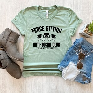 May include: A light green t-shirt with the text "Fence Sitting Anti-Social Club Please Sit Over There" and a graphic of three chairs with a heart in the middle.