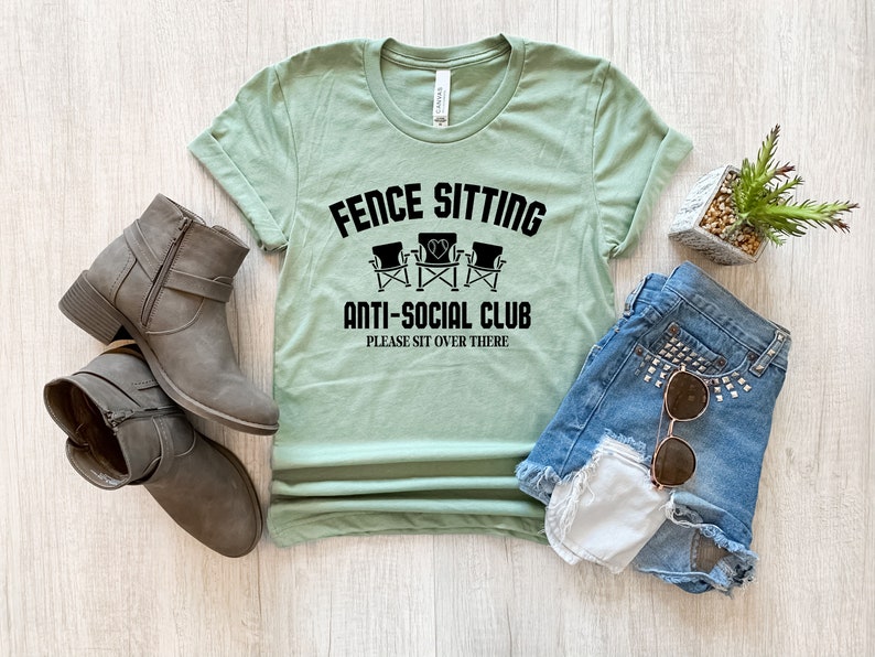 May include: A light green t-shirt with the text "Fence Sitting Anti-Social Club Please Sit Over There" and a graphic of three chairs with a heart in the middle.