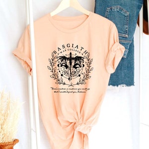 May include: A peach-colored t-shirt with a black graphic design featuring a skull with wings and the text "BASGIATH WAR COLLEGE" and a quote below.