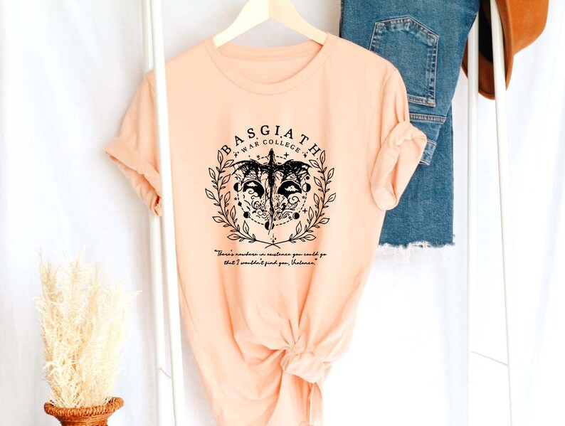 May include: A peach-colored t-shirt with a black graphic design featuring a skull with wings and the text "BASGIATH WAR COLLEGE" and a quote below.