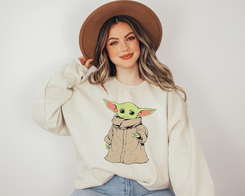 May include: A cream colored sweatshirt with a green cartoon character wearing a brown coat. The character has large ears and is looking to the left. The sweatshirt has a brown hat on top of it.