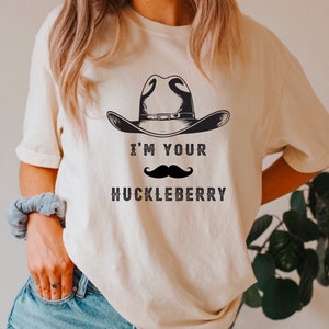 Puede incluir: Camiseta color crema con un sombrero de vaquero negro y un bigote. El texto "I'M YOUR HUCKLEBERRY" est&aacute; impreso debajo en una fuente vintage. La camiseta es de estilo casual y unisex.