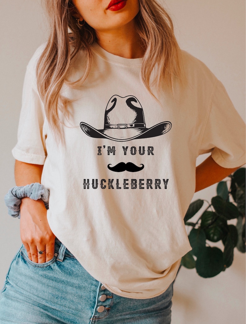 Puede incluir: Camiseta color crema con un sombrero de vaquero negro y un bigote. El texto "I'M YOUR HUCKLEBERRY" est&aacute; impreso debajo en una fuente vintage. La camiseta es de estilo casual y unisex.