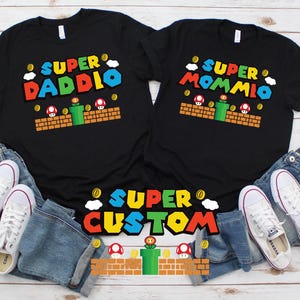 May include: Black t-shirts with colorful Super Mario-themed graphics. One shirt says "Super Daddio" and the other "Super Mommio". A third shirt says "Super Custom". White sneakers and blue jeans are also in the image.