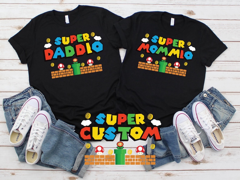 May include: Black t-shirts with colorful Super Mario-themed graphics. One shirt says "Super Daddio" and the other "Super Mommio". A third shirt says "Super Custom". White sneakers and blue jeans are also in the image.