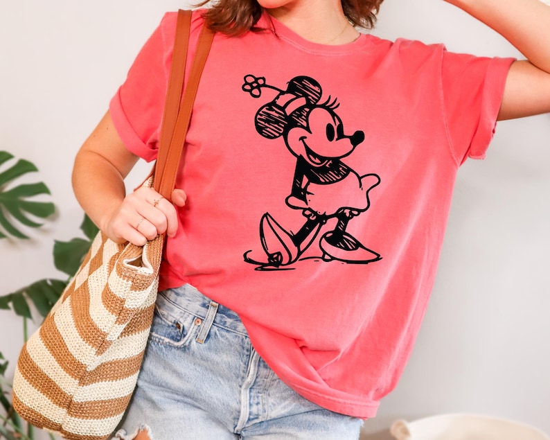 May include: A coral-colored t-shirt featuring a black graphic of Minnie Mouse. The cartoon character is depicted in a classic pose, wearing a skirt and shoes. The shirt has short sleeves and a relaxed fit, suitable for casual wear.