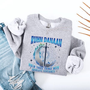 May include: Gray sweatshirt with a blue and white graphic design featuring a sword and a crescent moon. The text "Ruhn Danaan knew three things with absolute certainty" is printed on the sweatshirt.
