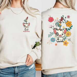 Puede incluir: Una sudadera beige con un dise&ntilde;o de dibujos animados de un cerdo y un gallo. El cerdo lleva una guirnalda de flores y el gallo sostiene una flor. El texto "MOANA" est&aacute; impreso debajo del dise&ntilde;o.