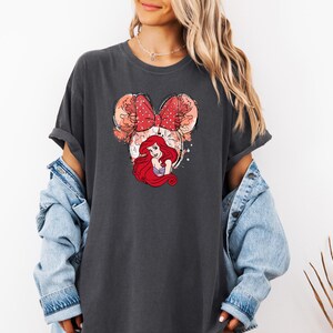 May include: A dark gray t-shirt with a graphic of Ariel from The Little Mermaid wearing a red bow. The graphic is in a cartoon style and has a watercolor effect.