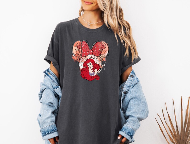 May include: A dark gray t-shirt with a graphic of Ariel from The Little Mermaid wearing a red bow. The graphic is in a cartoon style and has a watercolor effect.