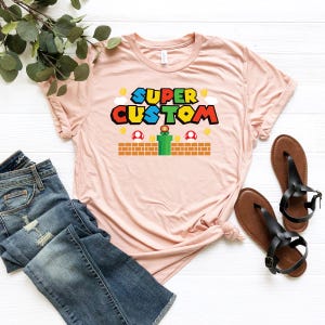 May include: A light pink t-shirt with the words "SUPER CUSTOM" in a colorful video game-themed design. The shirt is paired with blue jeans and black sandals. The design includes mushrooms, coins, and a green pipe.