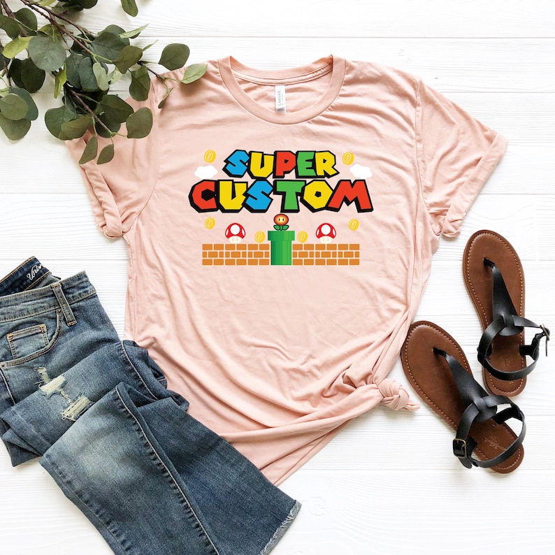 May include: A light pink t-shirt with the words "SUPER CUSTOM" in a colorful video game-themed design. The shirt is paired with blue jeans and black sandals. The design includes mushrooms, coins, and a green pipe.