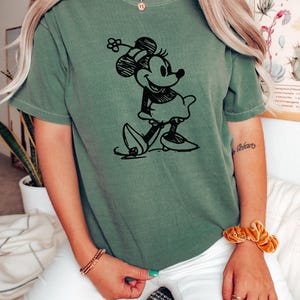 May include: Olive green t-shirt featuring a black sketch of Minnie Mouse. The graphic shows Minnie in a classic pose, wearing a dress and bow. The shirt is a relaxed fit.