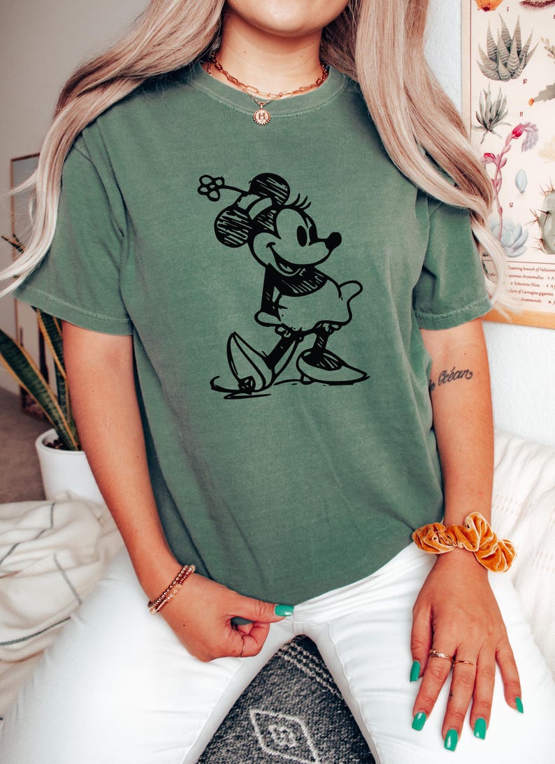 May include: Olive green t-shirt featuring a black sketch of Minnie Mouse. The graphic shows Minnie in a classic pose, wearing a dress and bow. The shirt is a relaxed fit.