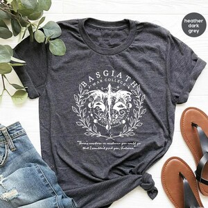May include: A heather dark grey t-shirt with a white graphic design featuring a winged creature, a circle with symbols, and the text "BASGIATH WAR COLLEGE". The text "There's nowhere in existence you could go that I wouldn't find you, holiness" is printed below the graphic.