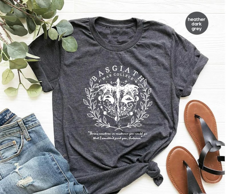May include: A heather dark grey t-shirt with a white graphic design featuring a winged creature, a circle with symbols, and the text "BASGIATH WAR COLLEGE". The text "There's nowhere in existence you could go that I wouldn't find you, holiness" is printed below the graphic.