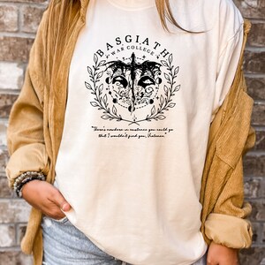 Peut inclure: T-shirt blanc avec un motif graphique noir repr&eacute;sentant une cr&eacute;ature ail&eacute;e, un cercle de lunes et le texte "BASGIATH WAR COLLEGE". La citation "There's nowhere in existence you could go that I wouldn't find you, Violanes." est imprim&eacute;e sous le graphique.