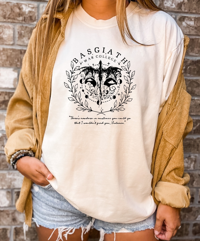 Peut inclure: T-shirt blanc avec un motif graphique noir repr&eacute;sentant une cr&eacute;ature ail&eacute;e, un cercle de lunes et le texte "BASGIATH WAR COLLEGE". La citation "There's nowhere in existence you could go that I wouldn't find you, Violanes." est imprim&eacute;e sous le graphique.